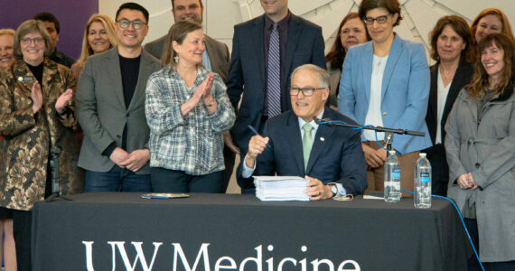 Washington Gov. Jay Inslee (seated) prepares to sign the 2024 supplemental budget on March 29, 2024. He was joined by several Democratic state lawmakers, including, to his right, Sen. June Robinson, the lead Senate budget writer. (Courtesy of UW Medicine | Nicholas Swatz)